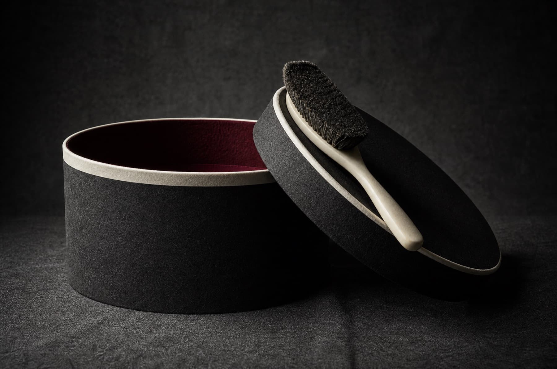 Open rigid charcoal hat box showing wine lining with a hat brush across the lid